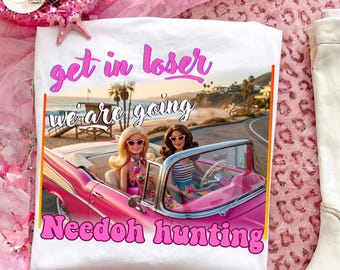 May include: A white t-shirt featuring a graphic of two dolls in a pink convertible car on a coastal road. The text reads "get in loser we are going Needoh hunting" in pink and white lettering. The design includes a beach scene with palm trees.