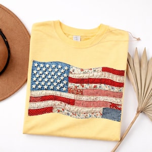 May include: Yellow t-shirt featuring a sewn-on American flag design. The flag has a blue denim field with white stars and red and white striped sections made of patterned fabric. A brown hat is visible in the background.