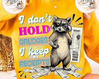 May include: A bright yellow sweatshirt featuring a graphic of a raccoon wearing black sunglasses. The raccoon holds a receipt, and the text reads: I don't HOLD GRUDGES I keep RECEIPTS. Additional receipts are scattered at the bottom of the design.
