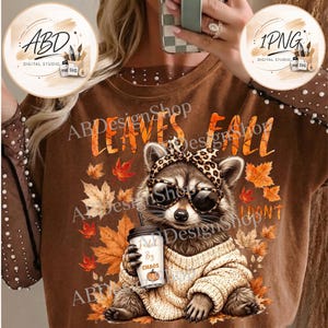 May include: A brown sweatshirt featuring a graphic of a raccoon wearing leopard print sunglasses and a matching headband, dressed in a beige sweater, holding a coffee cup labeled FUELED BY CHAOS with a pumpkin icon, surrounded by autumn leaves.