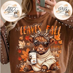 May include: A brown sweatshirt featuring a graphic of a highland cow wearing a leopard print headband and sunglasses, dressed in a cream sweater, holding a coffee cup labeled Fueled By Chaos, surrounded by autumn leaves and the text LEAVES FALL.