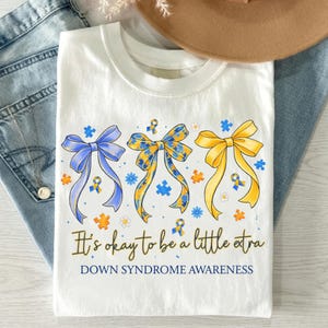 May include: White t-shirt with three colorful bows in blue, yellow, and patterned yellow and blue. The bows have ribbons and puzzle pieces, with the text "It's okay to be a little extra" and "Down Syndrome Awareness" below the bows.
