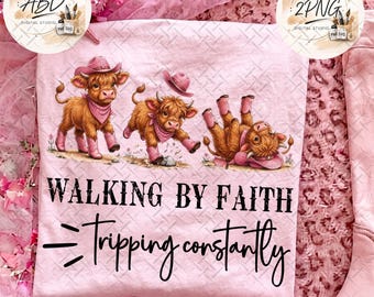 May include: A light pink t-shirt featuring three brown Highland cattle wearing pink cowboy hats and boots. The text reads WALKING BY FAITH and Tripping constantly in black lettering. The design includes small pink flowers on the ground.