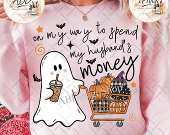 May include: A light pink sweatshirt featuring a white ghost holding a coffee cup and pushing an orange shopping cart filled with pumpkins. The text reads, on my way to spend my husband's money, accompanied by small black bats, orange stars, and swirls.