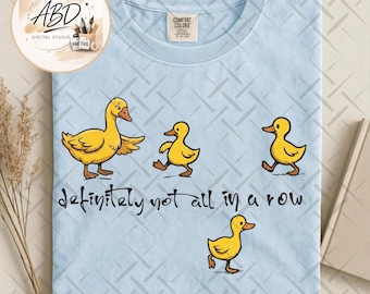 May include: A light blue t-shirt featuring a graphic of one adult duck and three small ducklings in yellow with orange beaks and feet. The text "definitely not all in a row" is printed in black script below the ducks. The shirt has a Comfort Colors label.