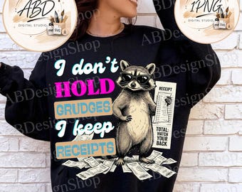 May include: A black long-sleeve sweatshirt featuring a graphic of a raccoon wearing sunglasses. The text reads I don't HOLD GRUDGES I keep RECEIPTS. The raccoon holds a receipt, and more receipts are scattered at its feet. The text TOTAL: WATCH YOUR BACK is printed on the receipt held by the raccoon.