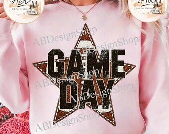 May include: A light pink crewneck sweatshirt featuring a large, distressed brown star graphic with a football texture pattern inside. The words "GAME DAY" are printed in bold, black, distressed capital letters across the centre of the star.