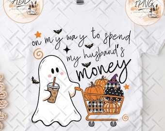 May include: A white t-shirt featuring a graphic of a ghost holding an iced coffee and pushing an orange shopping cart filled with pumpkins. The text reads, on my way to spend my husband's money, surrounded by small bats, stars, and swirl icons.
