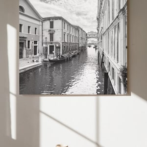 Peut inclure: Photographie en noir et blanc d'un canal à Venise, Italie. Des bâtiments bordent le cours d'eau, avec un pont au loin. Une gondole est amarrée le long du canal. L'image est affichée sur un mur.