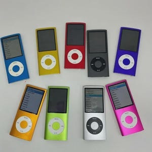 May include: A collection of nine rectangular digital music players arranged in two rows. The devices feature vertical screens and circular click wheels in various colors including blue, yellow, red, dark gray, purple, orange, green, silver, and pink.
