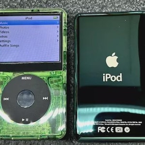 May include: Two Apple iPod digital media players. The left device features a translucent green casing, a black circular click wheel, and a screen displaying a menu with Music, Photos, Videos, Extras, Settings, and Shuffle Songs. The right device is black.