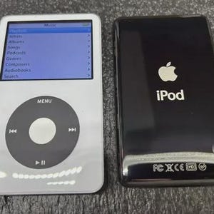 May include: A white digital music player with a circular navigation wheel and a screen displaying a music menu, positioned next to a black digital music player showing the Apple logo, the word iPod, and various regulatory certification symbols.