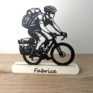 Könnte beinhalten: Schwarze Metallskulptur eines Radfahrers mit Rucksack und Packtaschen auf einem Fahrrad. Die Skulptur ist auf einem weißen Sockel mit dem Namen "Fabrice" in schwarzer Schrift montiert.