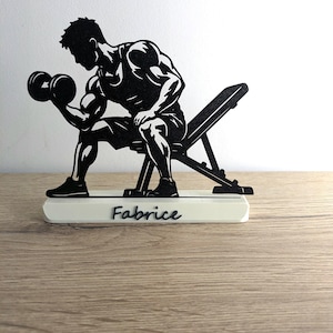 Könnte beinhalten: Schwarze Silhouette eines Gewichthebers, der auf einer Bank sitzt und Hanteln stemmt. Die Figur befindet sich auf einem weißen Sockel mit dem Namen "Fabrice" in Schreibschrift. Der Hintergrund ist eine helle Holzoberfläche.