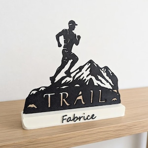 Peut inclure: Sculpture en bois noir et blanc représentant un coureur de trail sur une montagne. Le mot "TRAIL" et le nom "Fabrice" sont inscrits sur la base. Posé sur une surface en bois.