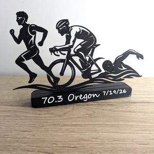 Peut inclure: Une sculpture en silhouette métallique noire représentant un triathlon avec un coureur, un cycliste et un nageur. Les figures sont montées sur une base rectangulaire noire avec le texte blanc 70.3 Oregon 7/19/26 imprimé sur la face avant.