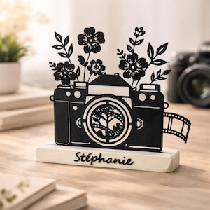 Könnte beinhalten: Schwarze dekorative Kamera-Silhouette mit floralen Akzenten und einem Filmstreifen-Detail. Die Kamera steht auf einem weißen Sockel mit dem Namen "Stéphanie" in Schrift. Auf einer Holzoberfläche.