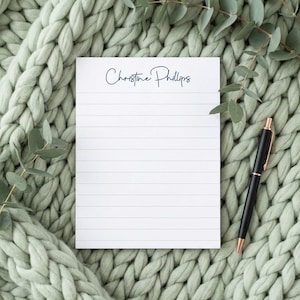 May include: A white lined notepad with the name "Christine Phillips" in cursive script, resting on a light green knitted blanket. A black and gold pen lies beside the notepad, with sprigs of greenery around the edges.
