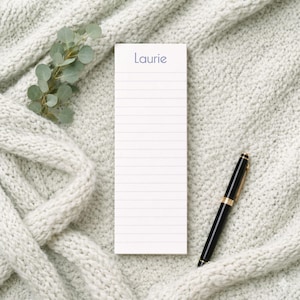 May include: A white lined notepad with the name "Laurie" printed in blue, a black pen with gold accents, and a sprig of eucalyptus. The notepad is on a light gray knitted blanket.