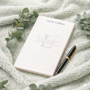 May include: A personalized notepad with lined paper, featuring the name "Laurie Collison" and a stylized "L" initial. A black and gold pen rests beside the notepad, all set against a soft, gray knitted blanket with eucalyptus sprigs.