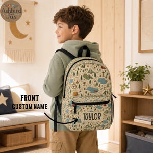 May include: A beige backpack featuring a repeating pattern of outdoor adventure icons including mountains, trees, compasses, and lakes. The front pocket displays the custom name "TAYLOR" in black lettering, with black straps and zippers.
