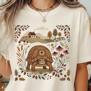 Könnte beinhalten: Ein cremefarbenes T-Shirt mit einem Grafikdruck einer Schildkröte mit komplexem Panzer-Muster. Das Design zeigt eine Landschaft mit einem Haus, sanften Hügeln, Bäumen, Weizenähren, Wildblumen und Eicheln in Braun-, Grün- und Rottönen.