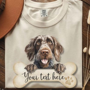 May include: A cream-colored t-shirt featuring a graphic of a brown dog with floppy ears and a happy expression. The dog rests its paws on a large, light-colored bone shape that contains the black text "Your text here" and a small paw print.