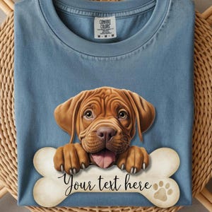 Könnte beinhalten: Ein blaues T-Shirt mit einer Grafik eines braunen Welpen, der seine Pfoten auf einen großen, hellen Knochen legt. Auf dem Knochen steht der Text "Your text here" in schwarzer Schreibschrift sowie ein kleiner Pfotenabdruck rechts.