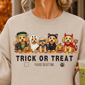 Può includere: Una felpa girocollo beige con una grafica di cinque cuccioli dorati in costumi di Halloween, tra cui Frankenstein, un fantasma, un supereroe, un gatto e un diavolo. Il testo recita TRICK OR TREAT PLEASE SELECT ONE con delle caselle.