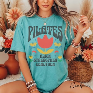 Può includere: T-shirt verde acqua con la scritta "PILATES" in grigio, una grafica di tulipano rosso e le parole "ALIGN STRENGTHEN LENGTHEN" in verde. La maglietta ha un design floreale retrò.
