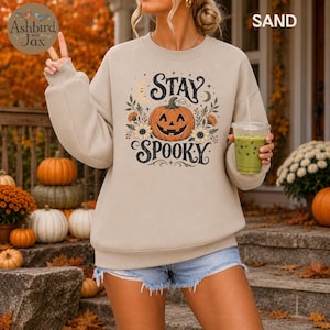 May include: A sand-colored crewneck sweatshirt featuring a graphic print of a jack-o'-lantern pumpkin surrounded by flowers, leaves, and a crescent moon. The text "STAY SPOOKY" is printed in a decorative black font above and below the pumpkin.