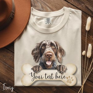 Peut inclure: T-shirt ivoire avec une illustration détaillée d'un chien brun et blanc tenant un os sur lequel est écrit "Your text here". Les pattes du chien reposent sur l'os. Le t-shirt a un col rond et la marque "Comfort Colors" est visible.