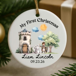 Könnte beinhalten: Ein rundes, weißes Keramikornament mit dem Text My First Christmas, Liam Lincoln und 09.23.26. Die Illustration zeigt einen Steinturm, einen kleinen Drachen, ein Kind als Ritter mit Schild und Schwert, einen grünen Baum, Sonne und Wolken.