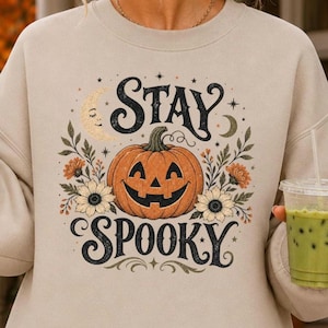 May include: A beige crewneck sweatshirt featuring a central graphic of a jack-o'-lantern pumpkin surrounded by sunflowers, leaves, and crescent moons. The text "STAY" appears above the pumpkin and "SPOOKY" below in a decorative black font.