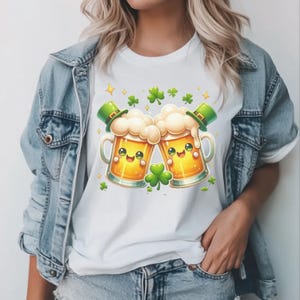 Peut inclure: T-shirt blanc avec un motif de dessin anim&eacute; de deux chopes de bi&egrave;re qui trinquent. Les chopes ont des visages souriants, de la mousse et portent des chapeaux de lutin verts. Des tr&egrave;fles verts et des &eacute;toiles dor&eacute;es sont dispers&eacute;s autour des chopes.