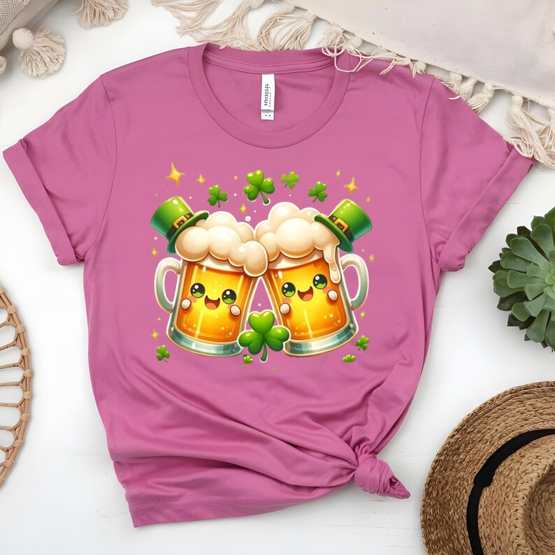 Peut inclure: T-shirt rose avec un motif de la Saint-Patrick. Le graphisme repr&eacute;sente deux chopes de bi&egrave;re de dessin anim&eacute; avec des visages souriants, de la mousse et des chapeaux de lutin verts. Des tr&egrave;fles verts et des &eacute;toiles dor&eacute;es sont dispers&eacute;s.