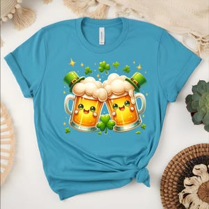 Peut inclure: Un t-shirt turquoise avec un motif de la Saint-Patrick. Le graphisme repr&eacute;sente deux chopes de bi&egrave;re de dessin anim&eacute; avec des visages souriants, de la mousse et des chapeaux de lutin verts. Des tr&egrave;fles verts et des &eacute;toiles dor&eacute;es sont dispers&eacute;s autour des chopes.