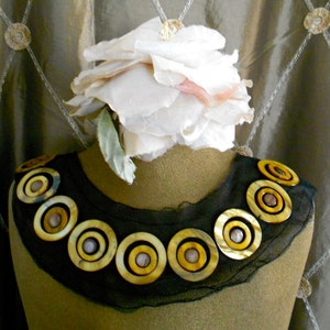 May include: A decorative necklace with large, circular, shell-like embellishments in shades of yellow and brown. The necklace is set on a black mesh base and displayed on a mannequin. A large white flower is placed above the necklace.