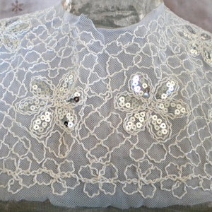 May include: A delicate, light blue lace collar with a floral design. The collar features a net-like pattern with embroidered flowers embellished with silver sequins. The collar is a vintage style.