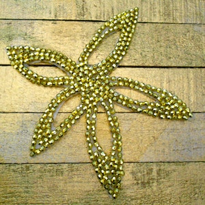 May include: A gold-colored, beaded floral applique with a five-petal design. The beads are densely packed, creating a textured surface. The applique is set against a gold-painted wooden background.