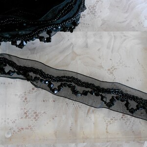 May include: Black beaded ribbon with a sheer, dark mesh base. The ribbon features rows of small, round beads and larger, faceted beads. The ribbon is partially rolled and displayed on a clear acrylic surface.