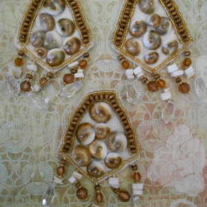 May include: Three decorative seashell ornaments. Each ornament features a white felt base, adorned with brown and white seashells, surrounded by a beaded border. Clear and brown crystal-like beads dangle from the bottom.