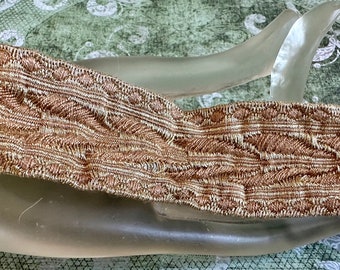 May include: A roll of decorative trim with a woven design in shades of brown and gold. The trim features a repeating pattern of leaves and is likely used for embellishing clothing or home decor items.