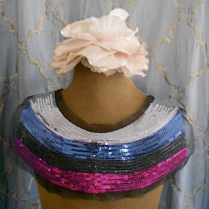 May include: A decorative collar with a layered design of sequins in silver, blue, black, and fuchsia. The collar is on a mannequin with a large, light pink flower on top.