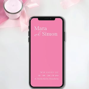 Könnte beinhalten: Ein Smartphone zeigt einen pinkfarbenen Bildschirm mit den Namen "Mara und Simon" in weißer Schrift. Der Text "WIR SAGEN JA!" mit Datum und Uhrzeit sowie der Ort "St. Martin Kirche, Rosenheim" sind ebenfalls zu sehen. Eine brennende Kerze in einem Glas und rosa Band im Hintergrund.