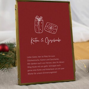 Könnte beinhalten: Rechteckiges, goldgerahmtes Schild mit tiefrotem Hintergrund. Es zeigt weiße Strichzeichnungen von zwei Geschenkpaketen und die Worte "Karten & Geschenke". Darunter befindet sich eine Nachricht auf Deutsch.