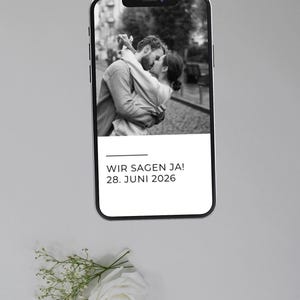 May include: A smartphone displays a black and white photo of a couple embracing and kissing. Below the photo, the text reads "WIR SAGEN JA! 28. JUNI 2026." A white rose and sprigs of baby's breath are at the bottom.