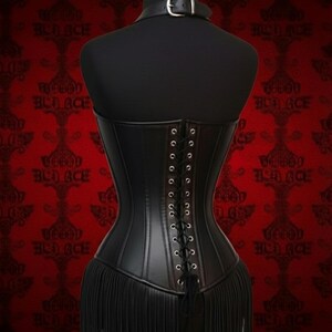 May include: Black corset with a fringe hem, displayed on a mannequin. The corset features a lace-up back and a small collar with a buckle. The background is red with a repeating pattern.