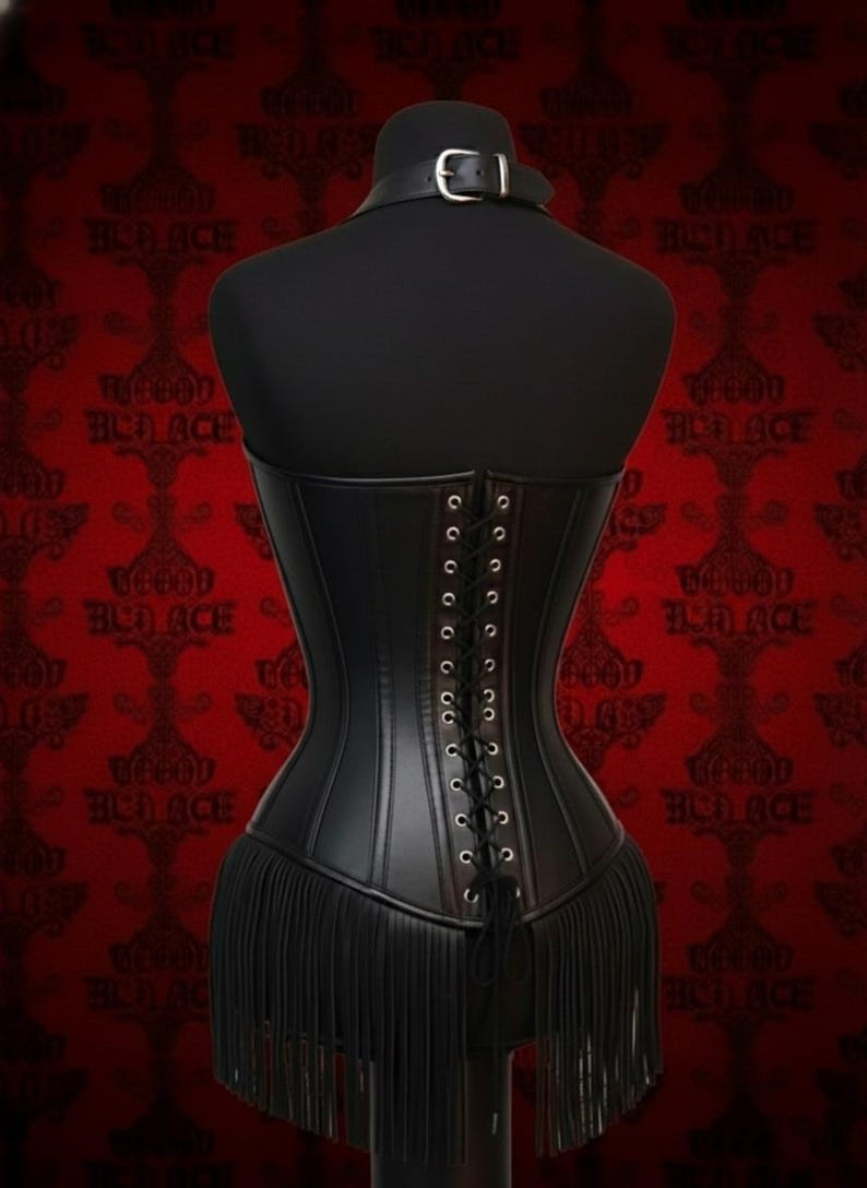 May include: Black corset with a fringe hem, displayed on a mannequin. The corset features a lace-up back and a small collar with a buckle. The background is red with a repeating pattern.