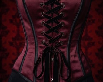 Puede incluir: Un corsé de satén color burdeos profundo con cordones de cinta de terciopelo negro a través de ojales de metal oscuro. La prenda incluye ribetes negros, un escote corazón con detalles de encaje negro y una larga cinta de terciopelo en la cintura.